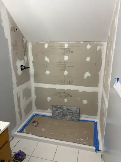 Shower alcove mid-remodel with cement backer board walls, taped seams, protected pan, and a tile sample.