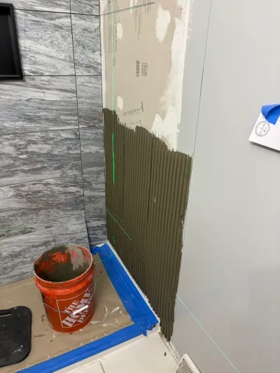 Bathroom corner mid-tile job: gray marble-look tile on one wall, combed thinset and laser lines on the next.