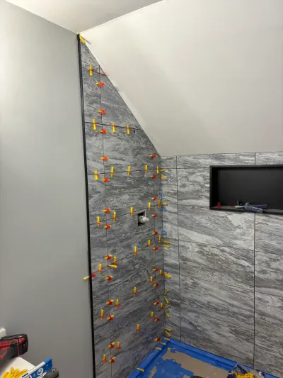 Shower walls mid-tiling with gray marble-look porcelain, leveling clips in place, under a sloped ceiling