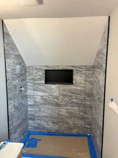 Shower alcove under a sloped ceiling tiled in large gray marble-look porcelain with a black-trimmed niche, mid-construction.