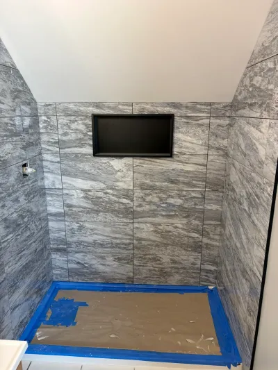 Gray marble-look tiled walk-in shower with black-framed niche under a sloped ceiling, floor masked off.
