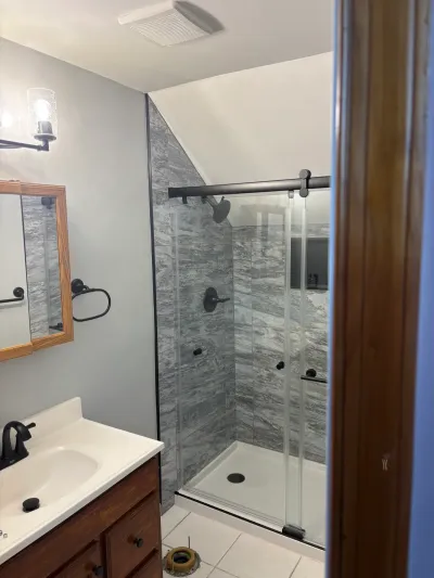 Remodeled bathroom with gray marble-look corner shower under a sloped ceiling and oak vanity with black fixtures.