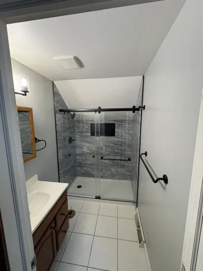 Narrow bathroom with gray marble-look shower, matte black sliding glass door, sloped ceiling and wood vanity.