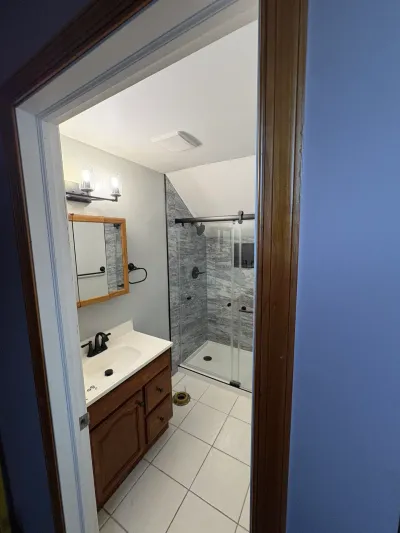 Small bathroom with oak vanity, gray marble-look corner shower and sloped ceiling, seen from the doorway.