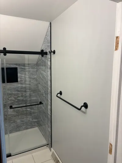 Corner glass shower with gray marble-look tile under a sloped ceiling, matte black hardware and towel bar.