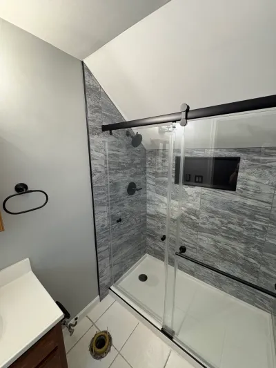 Gray tiled sloped-ceiling shower with matte black sliding glass door; toilet flange still open on white tile floor.