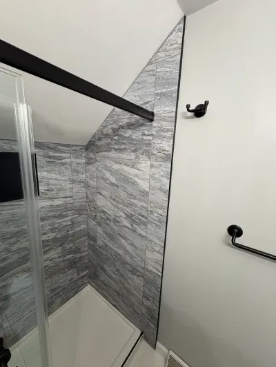 Gray marble-look tiled shower under a sloped ceiling with matte black curtain rod, hook and towel bar.