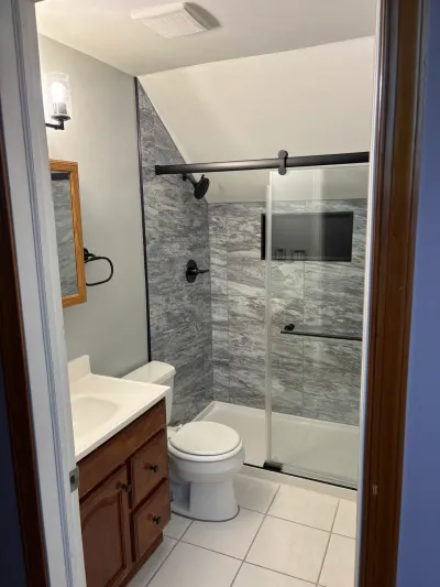 Remodeled small bathroom with gray tiled sloped-ceiling shower, oak vanity and white tile floor.