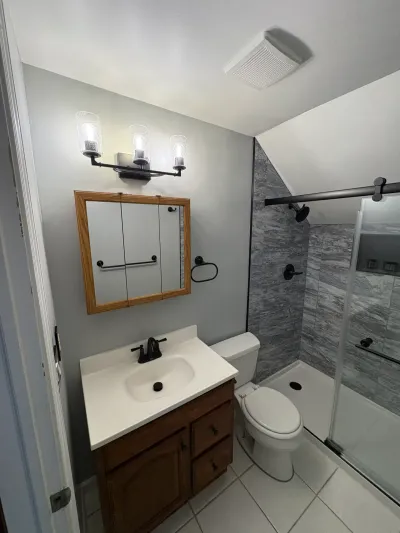 Small bathroom with oak vanity, gray marble-look tiled shower under a sloped ceiling, and matte black fixtures.