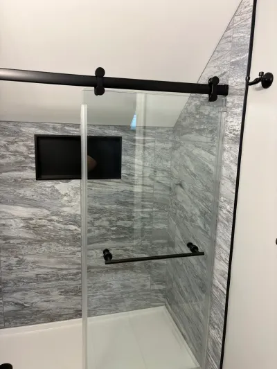 Walk-in shower with gray marble-look tile, sloped ceiling, matte black sliding glass door and recessed niche.