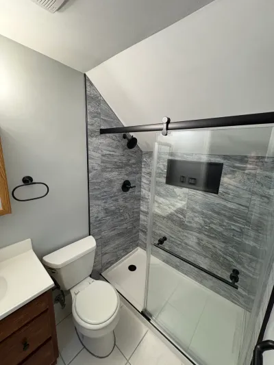 Attic bathroom with sloped ceiling, gray tile walk-in shower, black sliding glass door, toilet and wood vanity.