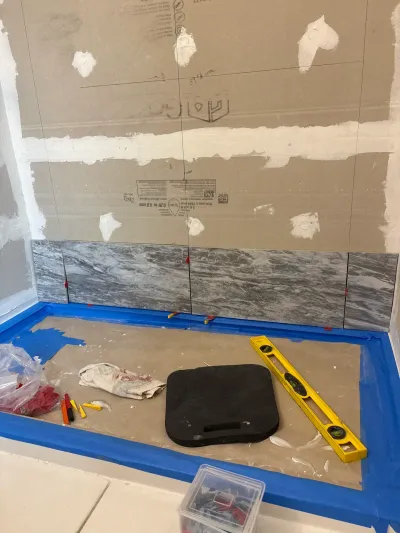 Shower walls in progress with backer board above a first row of gray marble-look tile and tools on the floor