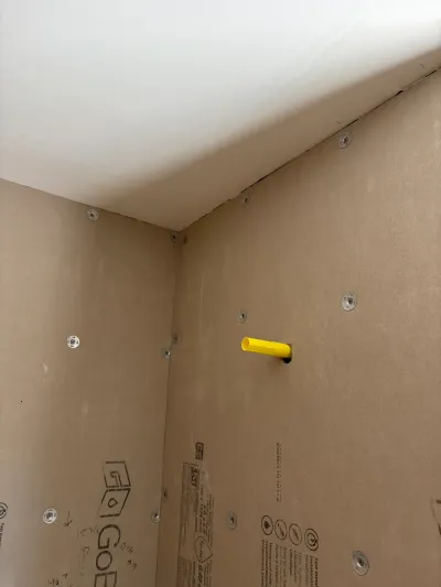 Bathroom alcove corner with tan backer board panels and a yellow plumbing stub-out awaiting tile.