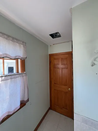 Narrow sage-green hallway with oak six-panel door, tile floor, curtained window, and an open ceiling light cutout.
