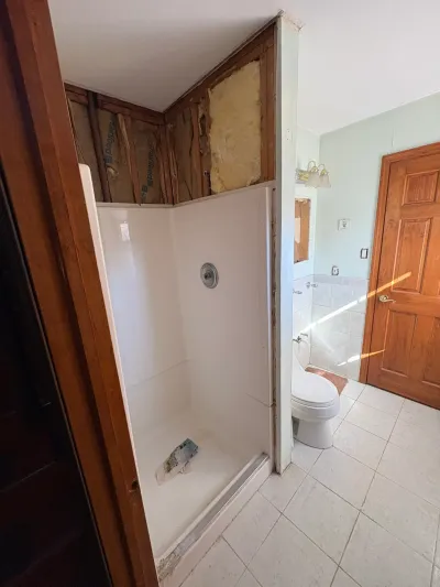 Bathroom mid-demolition with white fiberglass corner shower and exposed studs and insulation above it.