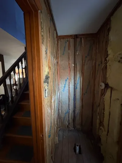 Narrow hallway stripped to studs with exposed insulation, bare plank floor, and staircase visible beyond wood casing.