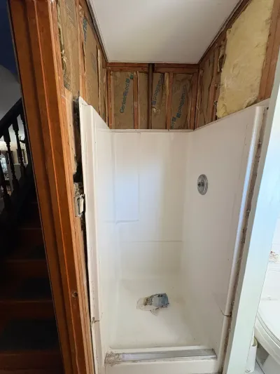 White fiberglass corner shower stall with drywall removed above, exposing studs and insulation before remodeling.
