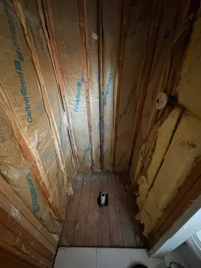 Narrow bathroom stripped to studs with kraft-faced insulation and a capped drain pipe in the subfloor.