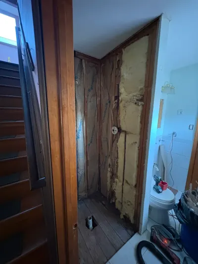 Bathroom shower alcove gutted to studs with exposed insulation, open drain in floor, and tools nearby.