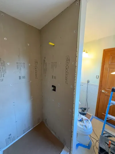 Corner shower alcove sheathed in tan GoBoard backer board during a bathroom remodel, ladder and tools nearby.