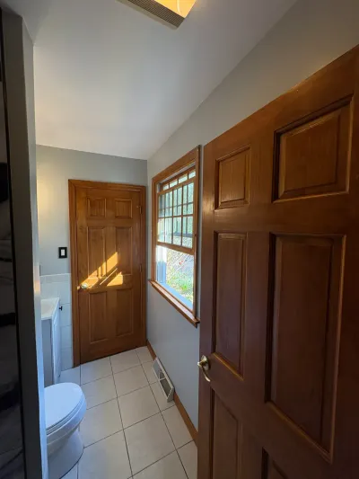 Narrow bathroom with diagonal beige tile floor, white tile wainscot, stained wood doors and a sloped ceiling.