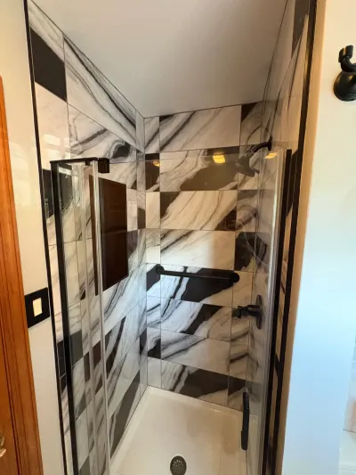 Narrow tiled shower with glossy marble-look tile, black accent stripe, matte black fixtures and glass door.