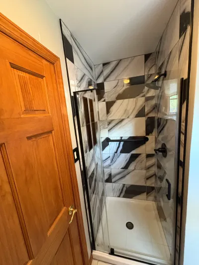 Walk-in shower with black-and-white marble-look tile, glass door, and matte black fixtures beside a wood door.