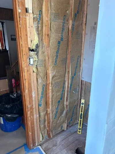 Opened wall framing with kraft-faced insulation, an outlet box, new drywall panel and a level during remodeling.