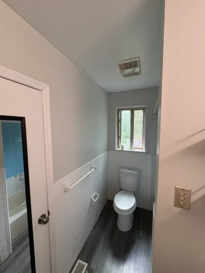 Narrow bathroom with white tile wainscot, gray walls, dark vinyl plank floor and toilet under a window.