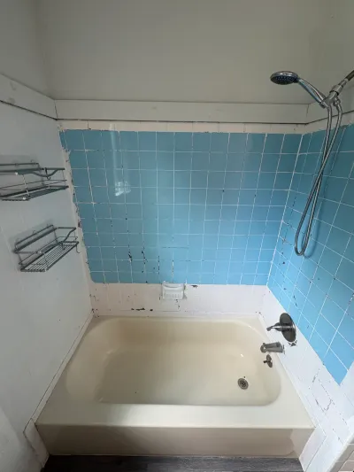 Dated bathroom tub alcove with chipped light blue tile, almond alcove tub, and chrome handheld shower.