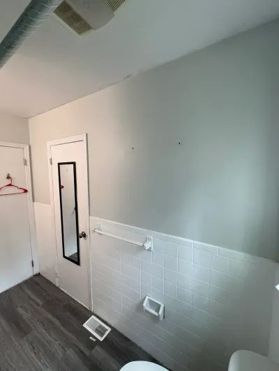 Existing bathroom corner with white tile wainscot, gray-green walls, mirrored door and gray plank flooring