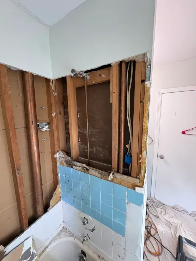 Bathroom demolition showing exposed studs and copper piping above a tub with blue and white tile