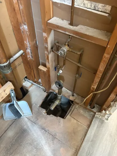 Bathroom wall opened to studs with copper pipes, brass shower valve, and cut subfloor exposing tub drain.