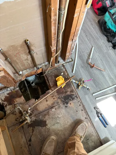 Bathroom floor opened up mid-remodel exposing copper and PEX plumbing, cast iron drain, and scattered tools.