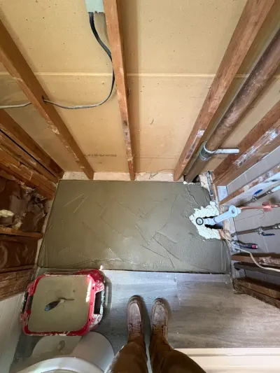 Overhead view of a bathroom floor mid-renovation with fresh gray mortar bed, exposed joists and plumbing rough-in.