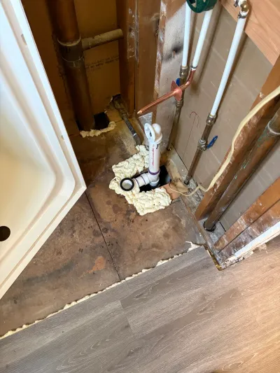 Bathroom corner mid-remodel with cut subfloor exposing shower drain plumbing and open stud wall pipes