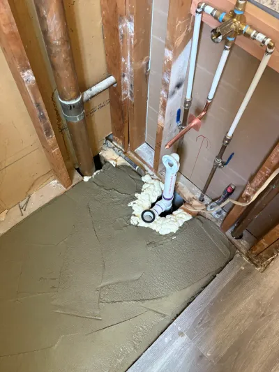 Shower corner mid-remodel with fresh mortar pan, exposed drain riser, and copper and PEX supply lines in open studs.