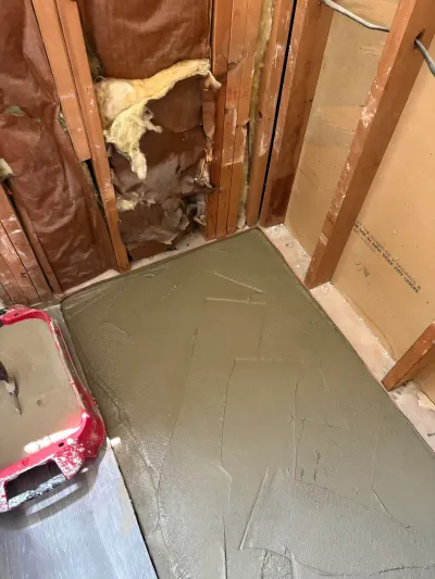 Freshly floated gray mortar shower pan in a corner bathroom alcove surrounded by open wood stud walls.