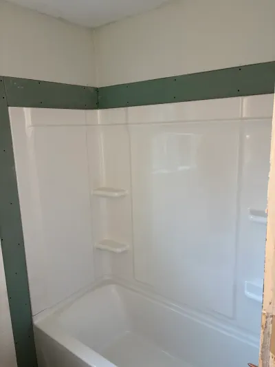 White tub surround newly installed over an alcove bathtub with unfinished green drywall above.