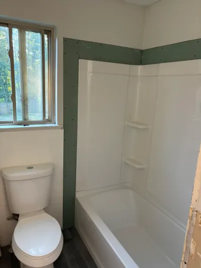Bathroom mid-remodel with new white tub surround, green drywall edges, toilet and window at left.