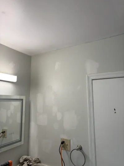 Bathroom wall covered in sanded spackle patches beside a mirror, vanity light, chrome towel ring and white door.