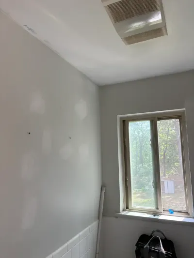 Bathroom corner mid-renovation with patched drywall, white tile wainscot, and a slider window.