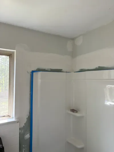 Bathroom under renovation with white corner shower surround, taped drywall patches, and a nearby window.