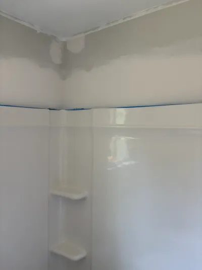 White fiberglass shower surround with corner shelves, blue painter's tape and patched drywall above.