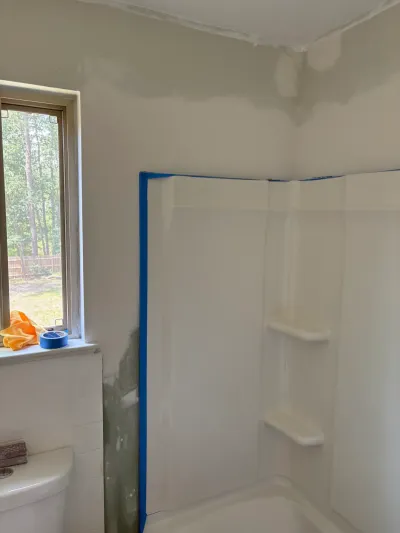 Bathroom mid-remodel with taped white tub surround, patched drywall, toilet and a sunlit window.