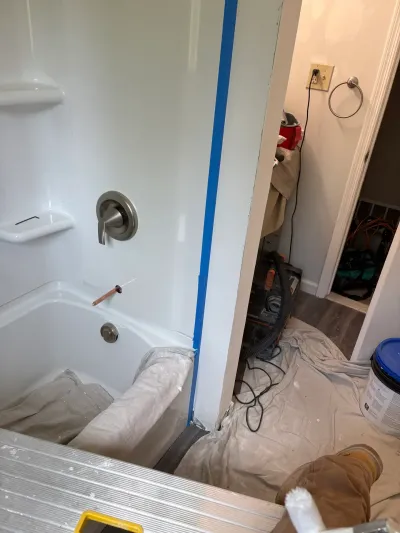 Bathroom mid-remodel with white tub surround, taped seams, drop cloths and a shop vac in the hallway.
