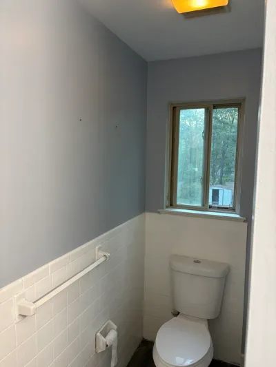 Narrow bathroom toilet alcove with white tile wainscot, gray-blue walls and a window above the toilet.