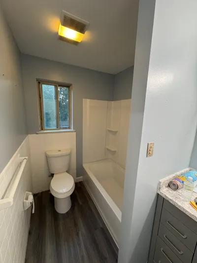 Narrow bathroom with white tub surround, tile wainscot, dark wood-look vinyl plank floor and gray vanity.