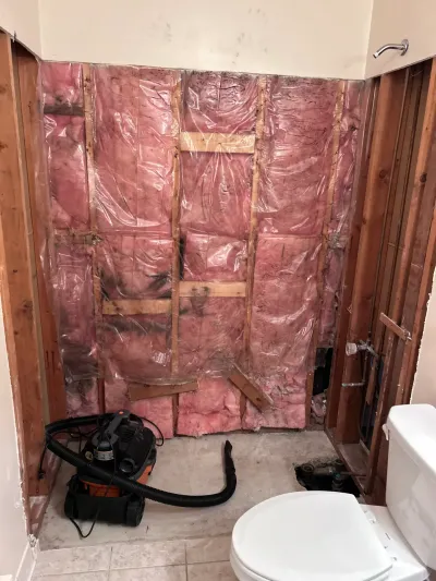 Bathroom tub alcove gutted to studs with pink insulation, vapor barrier, shop vac and remaining toilet.