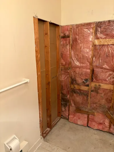 Bathroom wall stripped to studs with pink insulation and plastic sheeting during demolition.